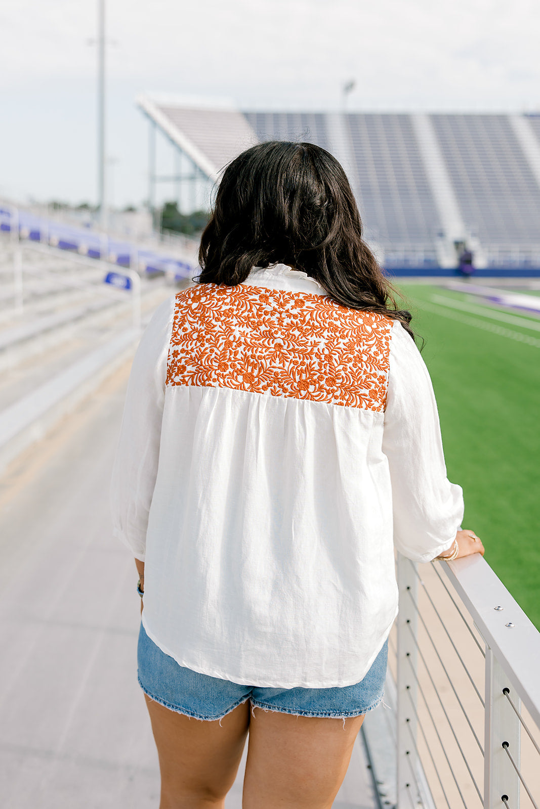 Woman with dark hair standing in a back profile position wearing the Jane Top at a football field. The focal point of this image is the pleating beneath embroidery and the burnt orange embroidery across shoulders. The model paired this top with light-wash denim shorts.
