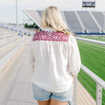 Woman with blonde hair standing in a back profile position wearing the Jane Top at a football field. The focal point of this image is the embroidery across the shoulders, pleating beneath, as well as emphasis on the ruffled collar. The model paired this top with light-wash denim shorts.