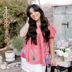 Woman in a colorful blouse and white shorts standing outdoors with a rustic background facing front. 