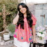 Woman in a colorful blouse and white shorts standing outdoors with a rustic background facing front. 