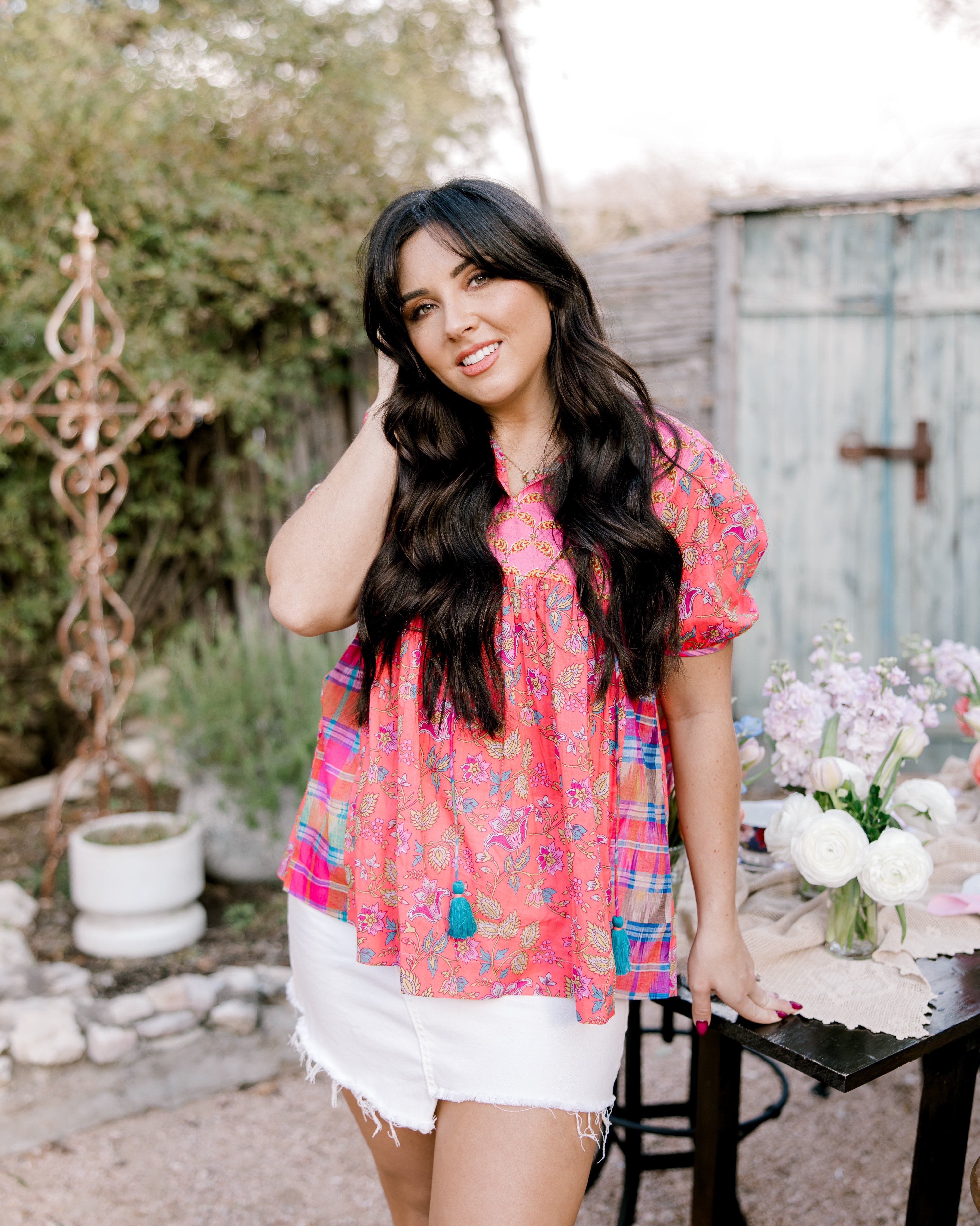 Woman in a colorful blouse and white shorts standing outdoors with a rustic background facing front. 