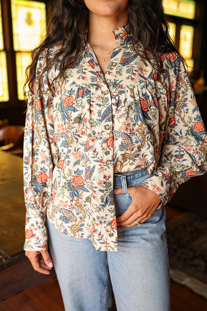 Woman with dark hair standing in a front profile position wearing the Jenna Top in a lifestyle scene. The focal point of this image is the buttons down front as well as pleating on chest and red/blue floral graphics. The model paired this top with light-wash denim jeans.
