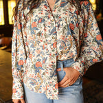 Woman with dark hair standing in a front profile position wearing the Jenna Top in a lifestyle scene. The focal point of this image is the buttons down front as well as pleating on chest and red/blue floral graphics. The model paired this top with light-wash denim jeans.