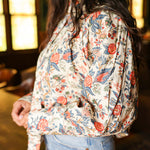 Woman with dark hair standing in a side profile position wearing the Jenna Top in a lifestyle scene. The focal point of this image is the red and blue floral pattern on sleeves as well as pleating. The model paired this top with light-wash denim jeans.