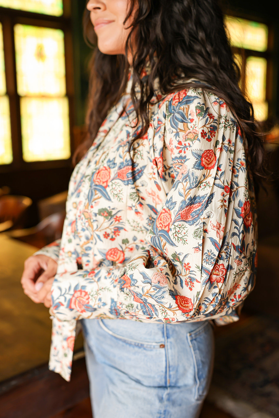 Woman with dark hair standing in a side profile position wearing the Jenna Top in a lifestyle scene. The focal point of this image is the red and blue floral pattern on sleeves as well as pleating. The model paired this top with light-wash denim jeans.