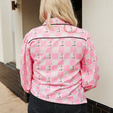 Woman with blonde hair wearing a button-down long-sleeve with black and pink boots graphics on a pink & white checkered background. the model paired this top with dark-wash denim jeans. 