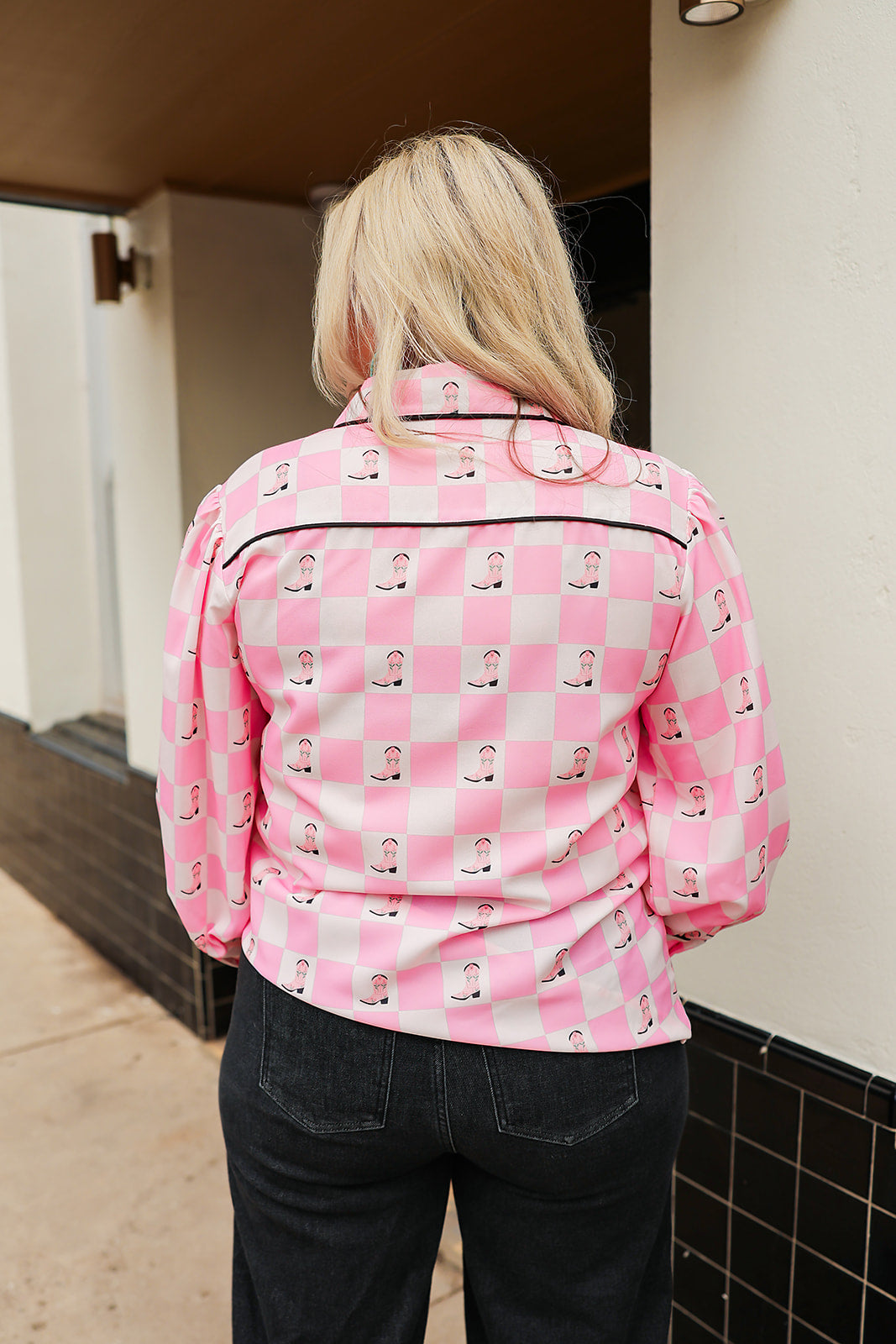 Woman with blonde hair wearing a button-down long-sleeve with black and pink boots graphics on a pink & white checkered background. the model paired this top with dark-wash denim jeans. 