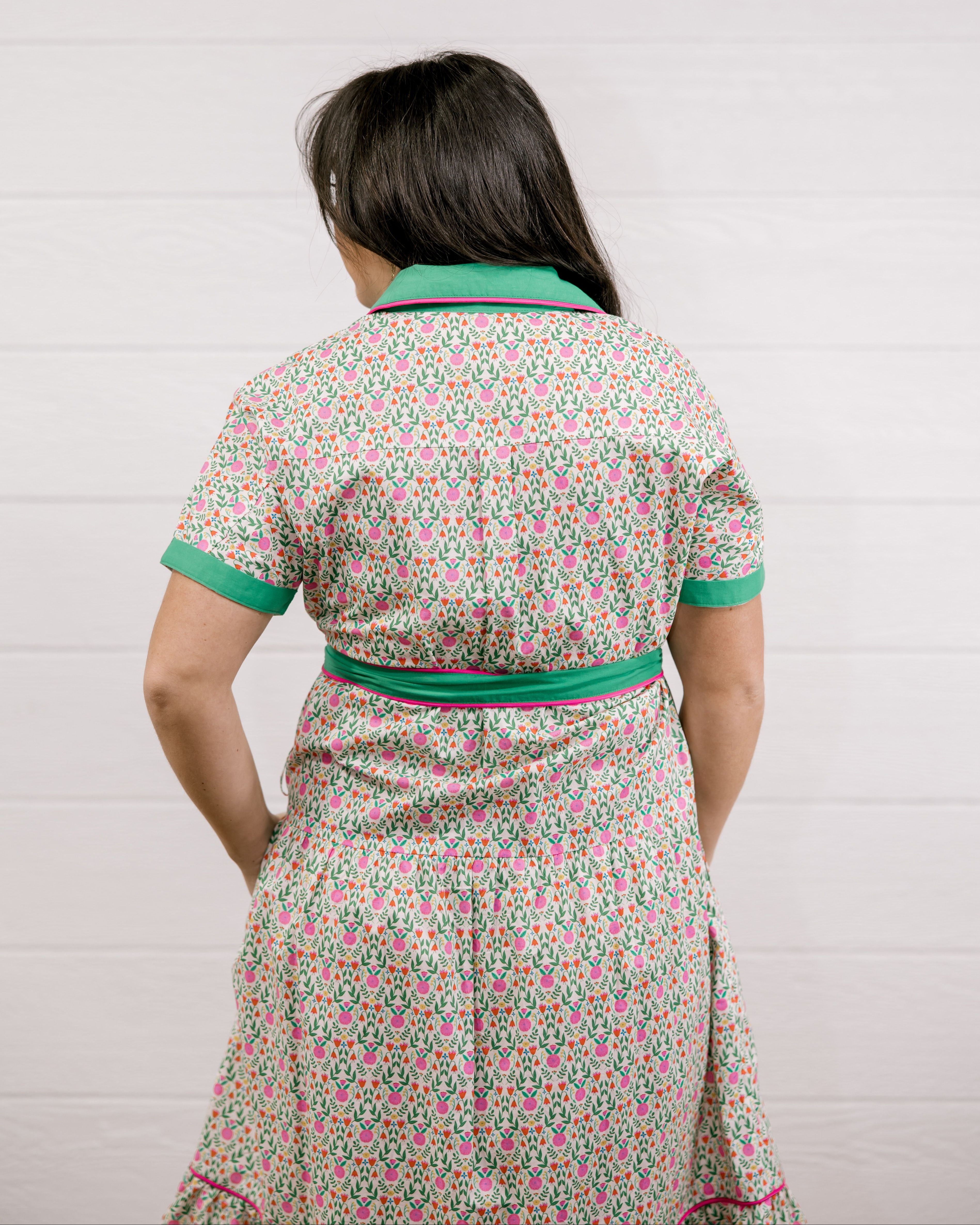 Lady wearing the a green and floral dress with pink piing and waist tie showing the back on a white background 