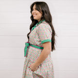 Lady wearing a green and floral dress with with pink piping showing the side and sleeve on a white background
