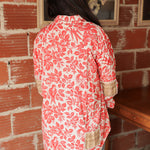 Woman with dark hair standing in a back profile position wearing the Karen Top against a red brick background. The focal point of this image is plaid patches on back and elbows of top as well as the red bird/floral graphics. The model paired this top with light-wash denim jeans. 
