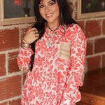 Woman with dark hair standing in a front profile position wearing the Karen Top against a red brick background. The focal point of this image is the red bird/floral graphics as well as buttons down the top and plaid patches on front and elbows. The model paired this top with light-wash denim jeans. 