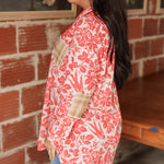 Woman with dark hair standing in a side profile position wearing the Karen Top against a red brick background. The focal point of this image is the red bird/floral graphics as well as the plaid patches of front and elbow of top. The model paired this top with light-wash denim jeans. 