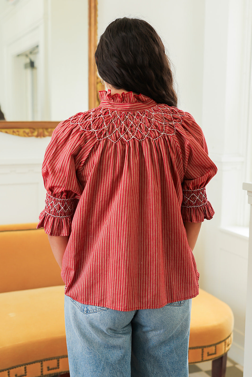 Woman with dark hair standing in a back profile position wearing the Kate Top in a lifestyle scene. The focal point of this image is the cream embroidery details on sleeve and back, ruffled collar, and striping pattern. The model paired this top with light-wash denim jeans. 
