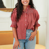 Woman with dark hair standing in a front profile position wearing the Kate Top in a lifestyle scene. The focal point of this image is the embroidery on the front as well as sleeves with with ruffles. The model paired this top with light-wash denim jeans. 