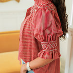 Woman with dark hair standing in a side profile position wearing the Kate Top in a lifestyle scene. The focal point of this image is the ruffled sleeves collar, and embroidery on chest and sleeve. The model paired this top with light-wash denim jeans. 