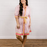 Woman wearing a patterned dress with a yellow belt against a white wooden panel background facing front. 