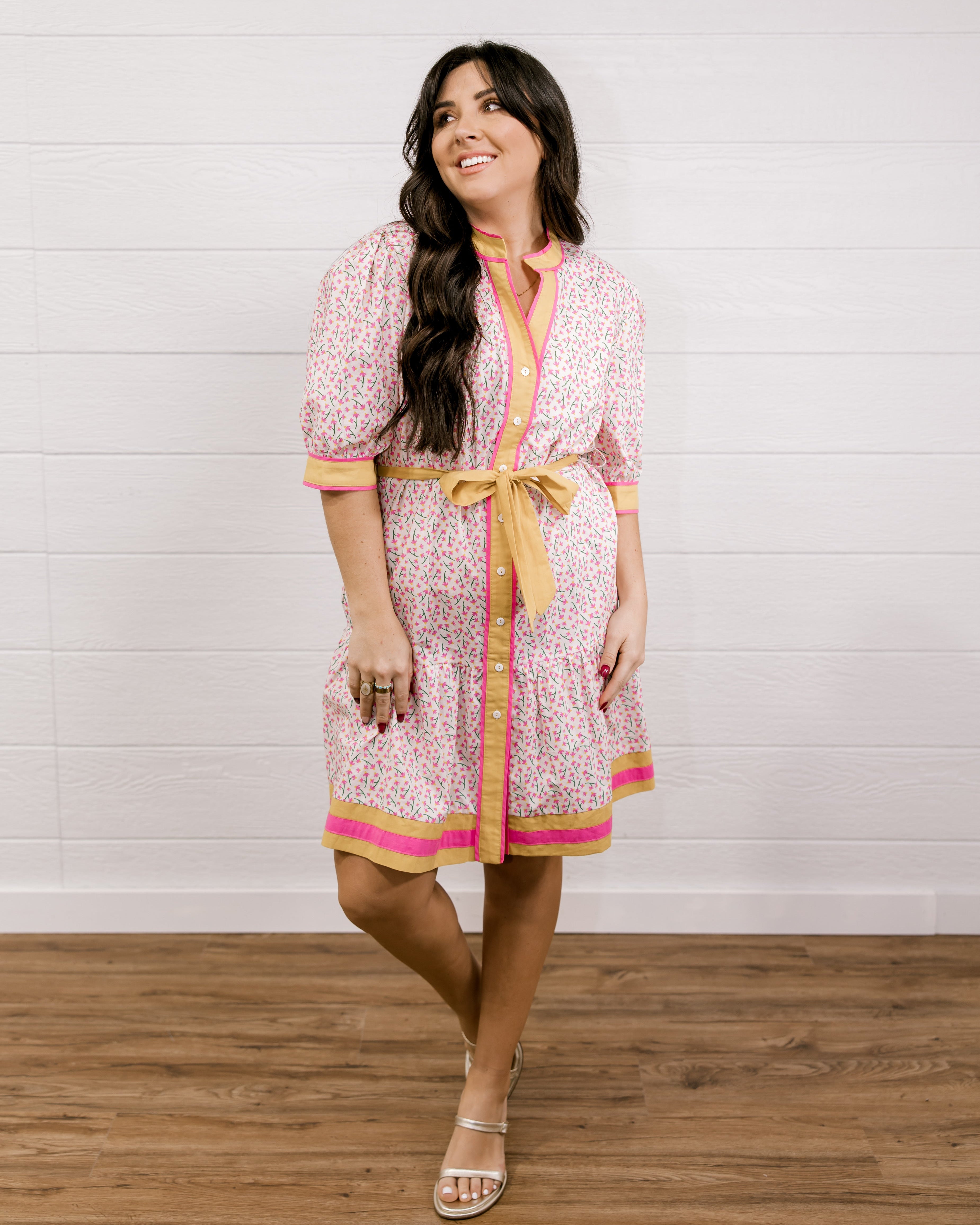 Woman wearing a patterned dress with a yellow belt against a white wooden panel background facing front. 