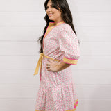 Woman wearing a pink floral dress with yellow accents against a white background facing side. 