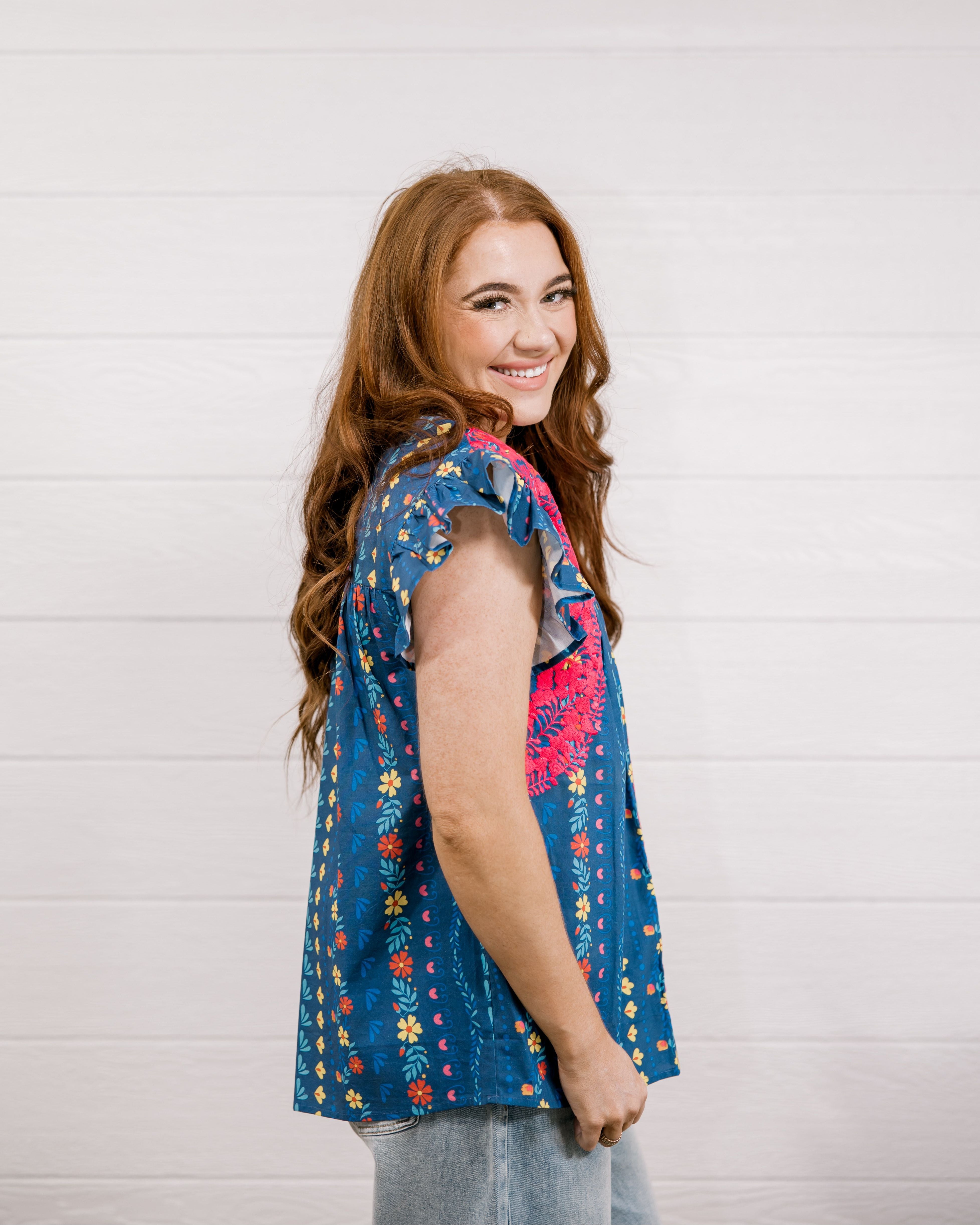 Women wearing a navy top with colorful patter, pink embroidery and ruffle sleeves  showing the side on a white background 