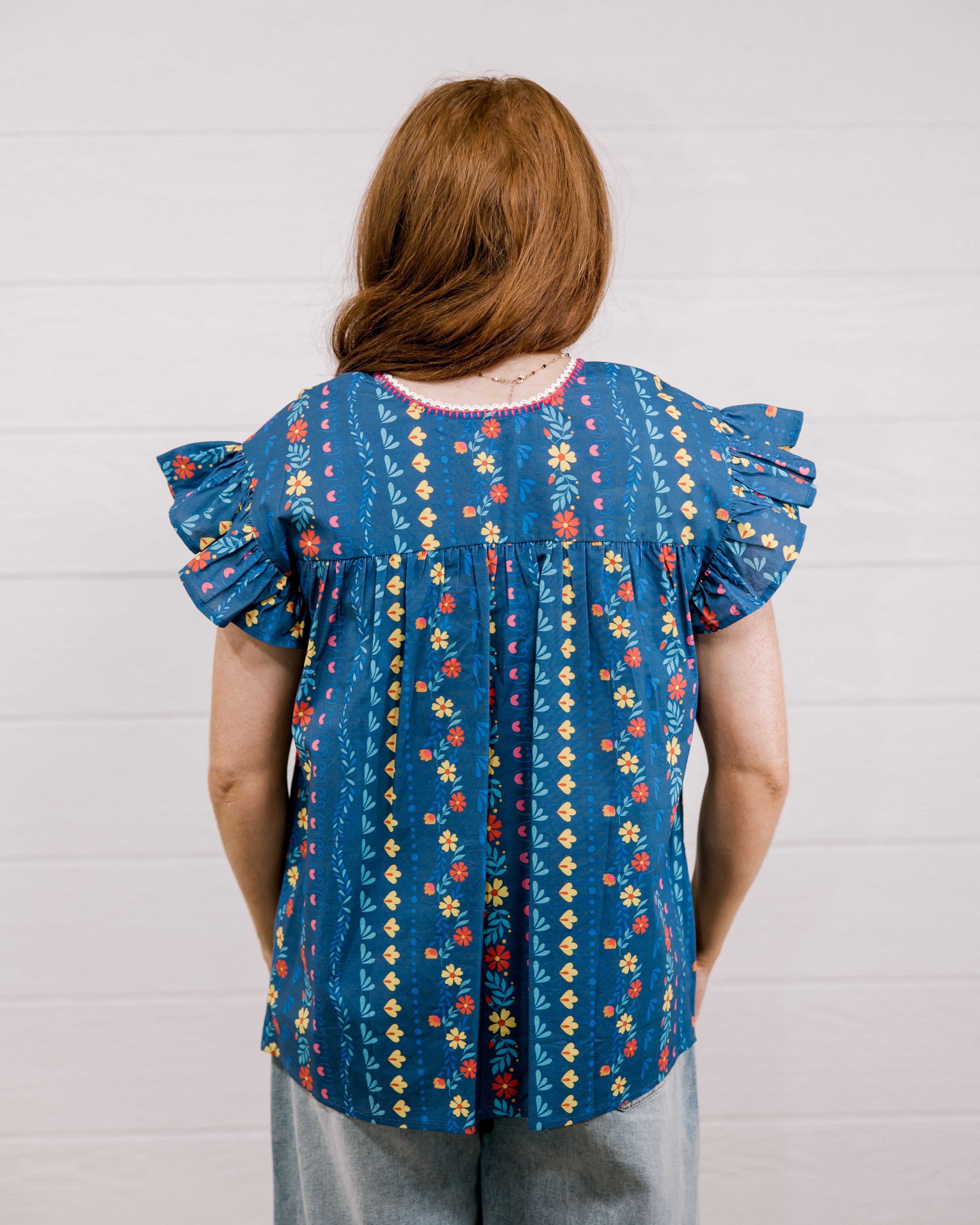 Women wearing a navy top with colorful patter, pink embroidery and ruffle sleeves  showing the back on a white background 