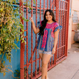 Women wearing a navy top with colorful patter, pink embroidery and ruffle sleeves in a colorful outdoor enviornment 
