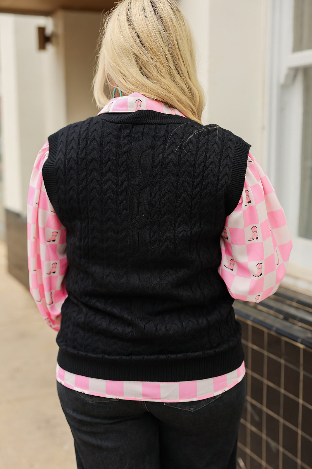 Woman with blonde hair standing in a back profile position wearing the Kodi Vest in a lifestyle scene. The focal point of this image is the cable knit pattern on back of sweater. The model paired this with dark-wash denim jeans and the Jesse Top.