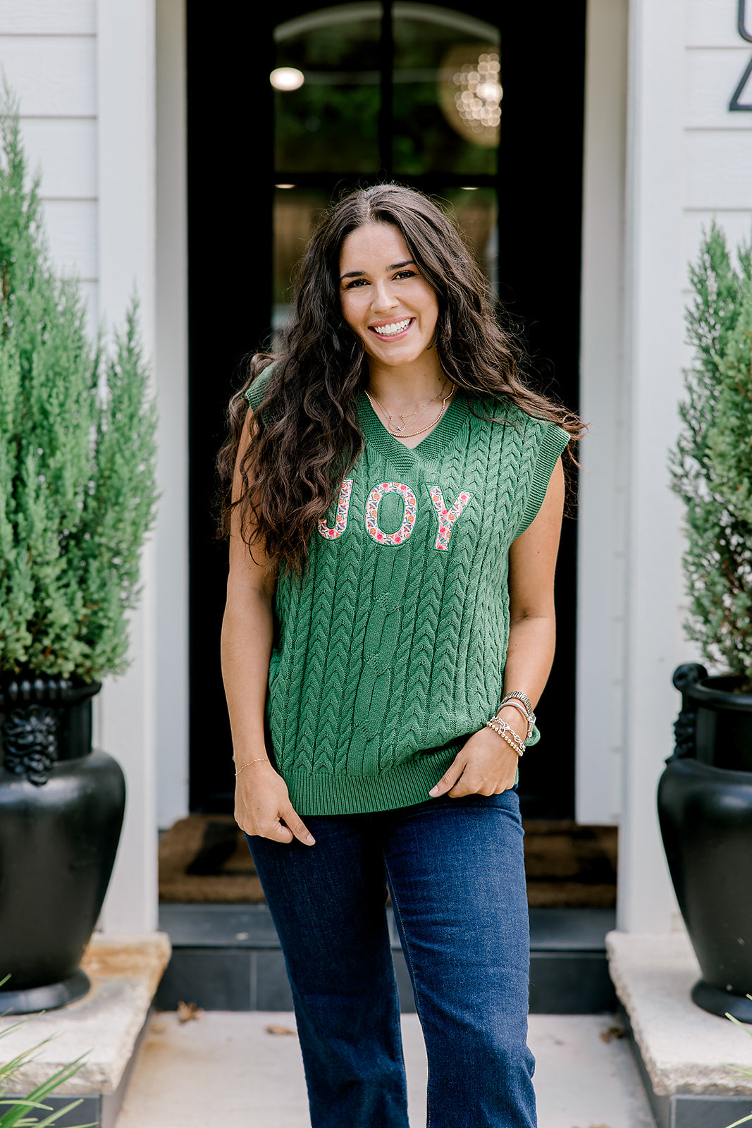 Woman with dark hair wearing a green sleeveless cable-knit vest with "JOY" embroidered on front. Model paired this top with dark-wash denim jeans. 