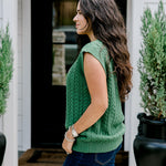 Woman with dark hair wearing a green sleeveless cable-knit vest with "JOY" embroidered on front. Model paired this top with dark-wash denim jeans. 