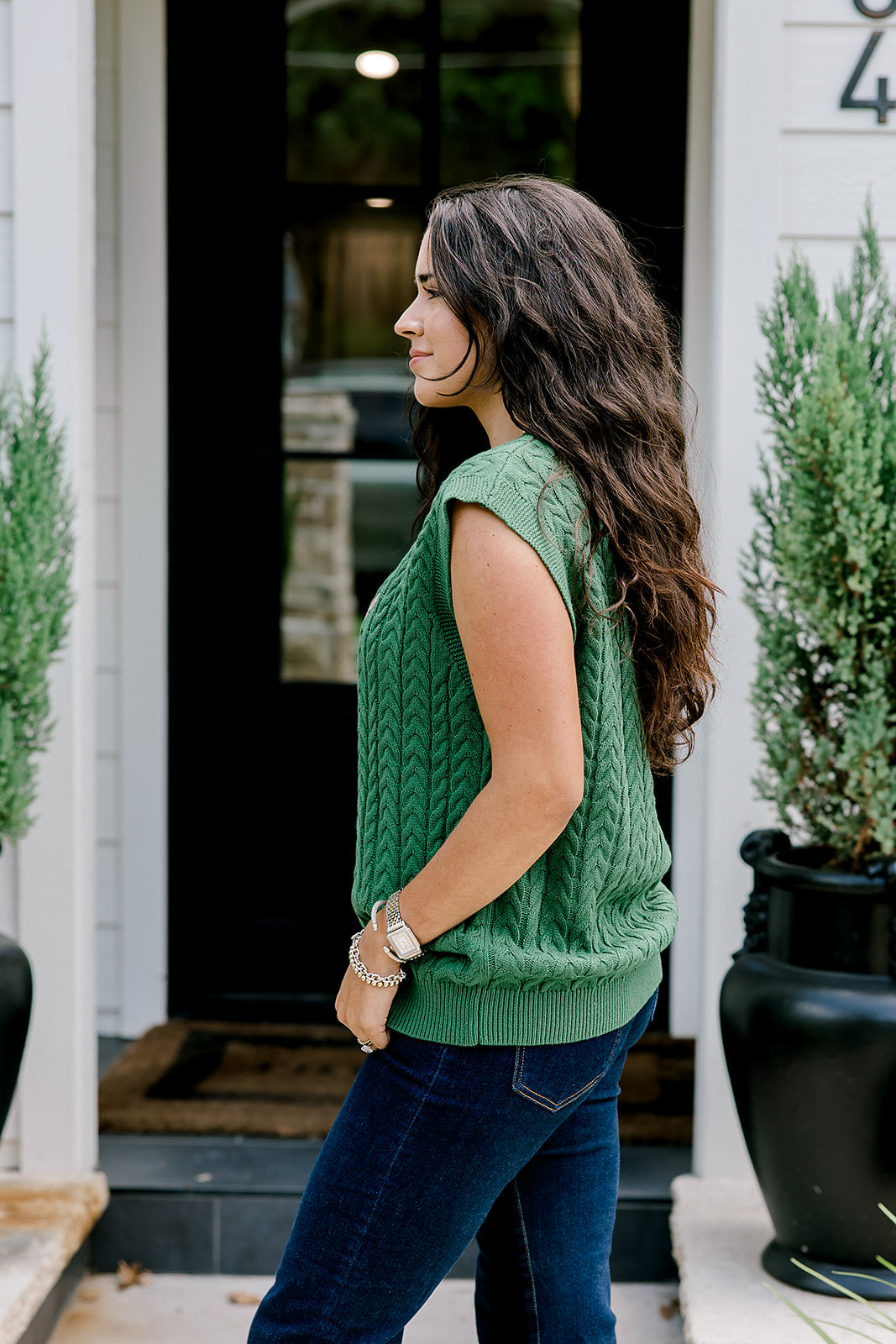 Woman with dark hair wearing a green sleeveless cable-knit vest with "JOY" embroidered on front. Model paired this top with dark-wash denim jeans. 