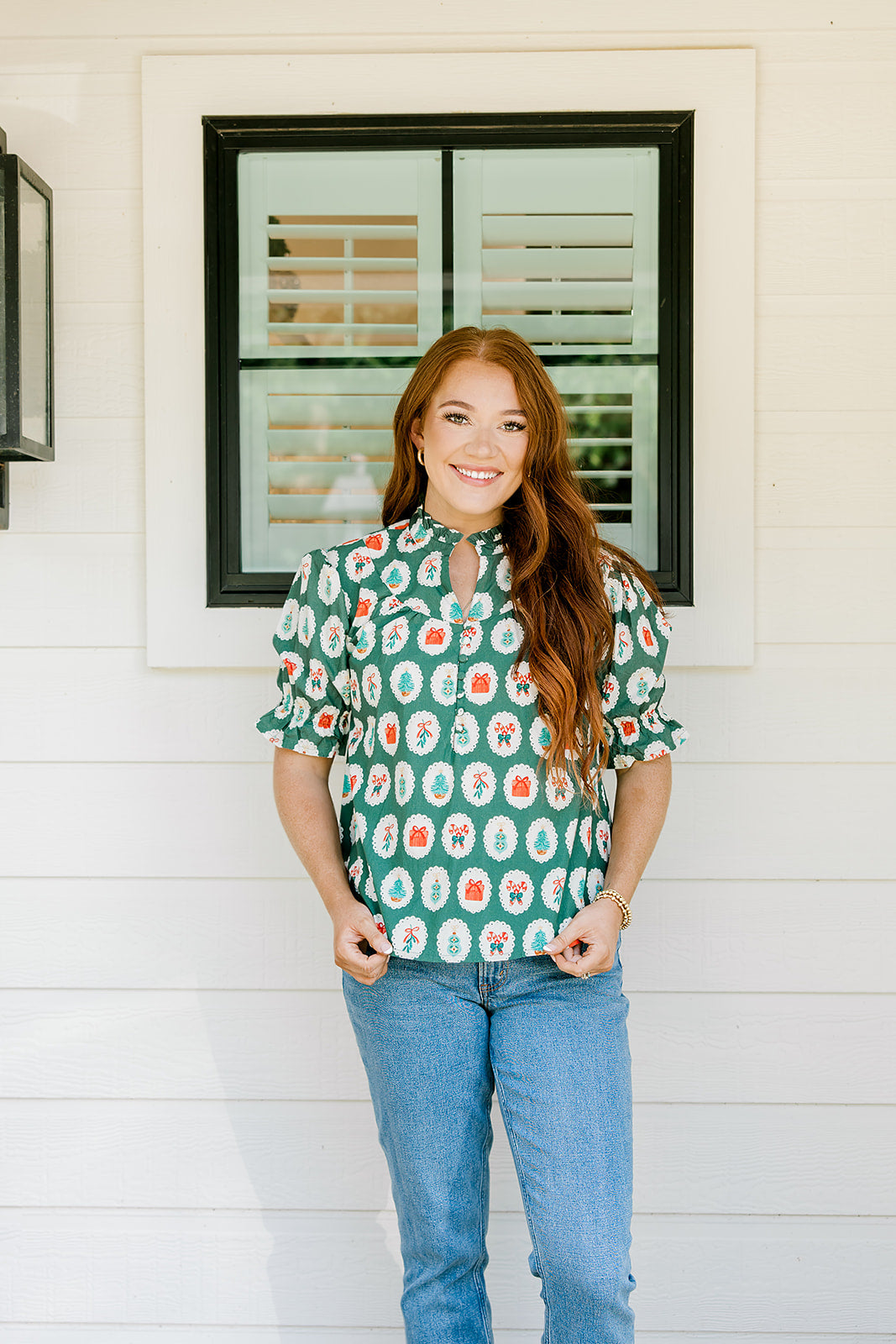 Woman with red hair wearing a short-sleeve top with Christmas item graphics, ruffled/pleated sleeves, and ruffled collar. Model paired this top with denim jeans. 