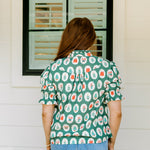 Woman with red hair wearing a short-sleeve top with Christmas item graphics, ruffled/pleated sleeves, and ruffled collar. Model paired this top with denim jeans. 