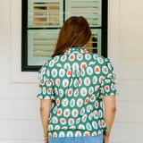Woman with red hair wearing a short-sleeve top with Christmas item graphics, ruffled/pleated sleeves, and ruffled collar. Model paired this top with denim jeans. 