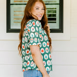 Woman with red hair wearing a short-sleeve top with Christmas item graphics, ruffled/pleated sleeves, and ruffled collar. Model paired this top with denim jeans. 