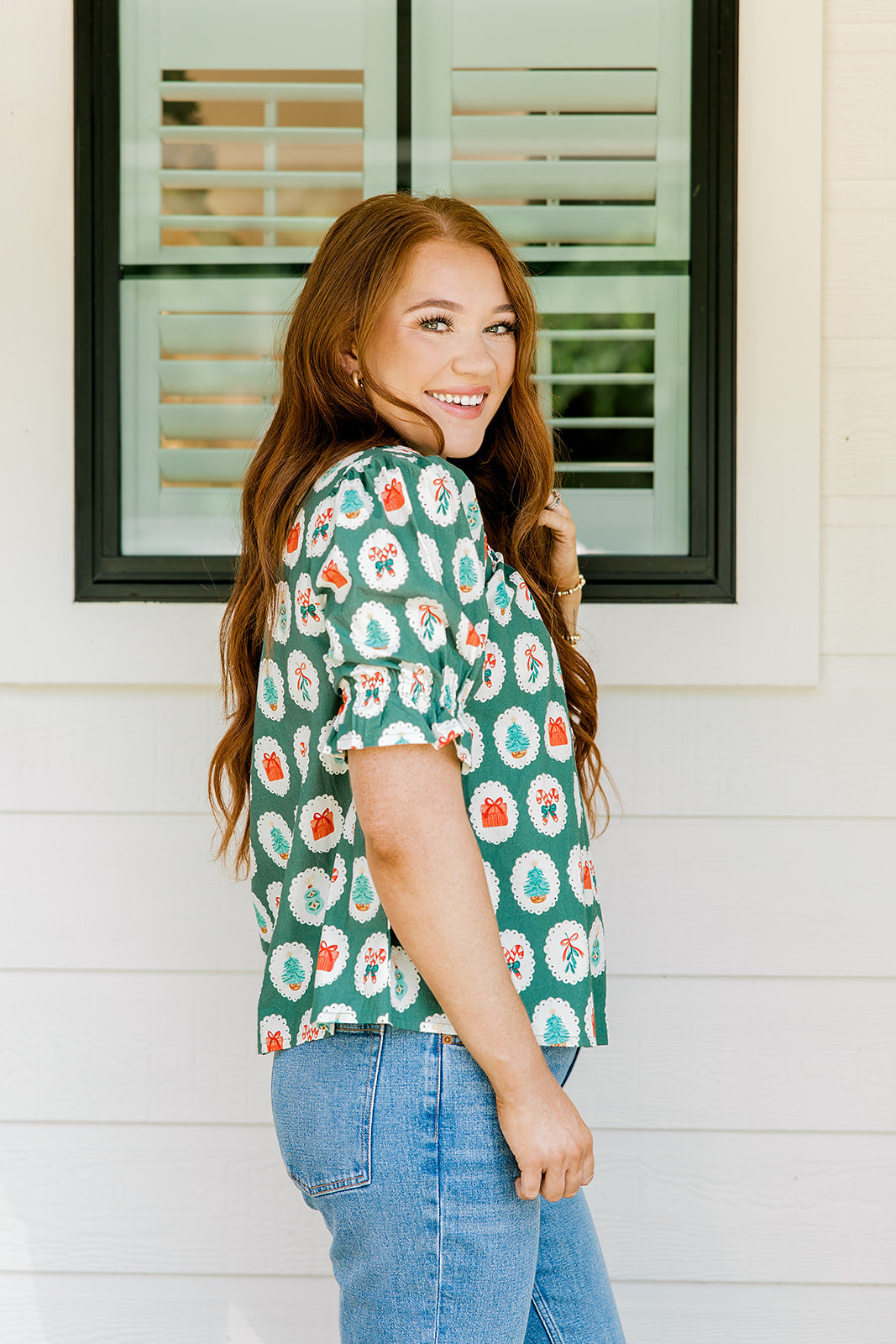 Woman with red hair wearing a short-sleeve top with Christmas item graphics, ruffled/pleated sleeves, and ruffled collar. Model paired this top with denim jeans. 