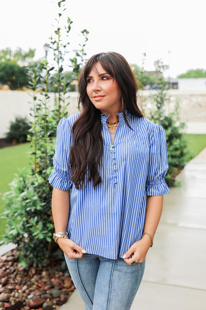 Woman with dark hair wearing a short-sleeve top with blue and white stripes, puffed sleeves, and buttons down front. Paired with light-wash denim jeans. 