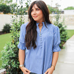 Woman with dark hair wearing a short-sleeve top with blue and white stripes, puffed sleeves, and buttons down front. Paired with light-wash denim jeans. 