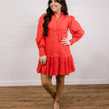 Woman wearing a red eyelet dress standing against a white wall.