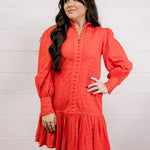 Woman wearing a red eyelet dress standing against a white wall.