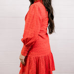 Side view of woman wearing a red eyelet dress standing against a white wall.