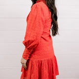 Side view of woman wearing a red eyelet dress standing against a white wall.