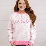Woman wearing a pink and white patterned sweatshirt with 'LOVED' embroidered in pink standing against a white background.