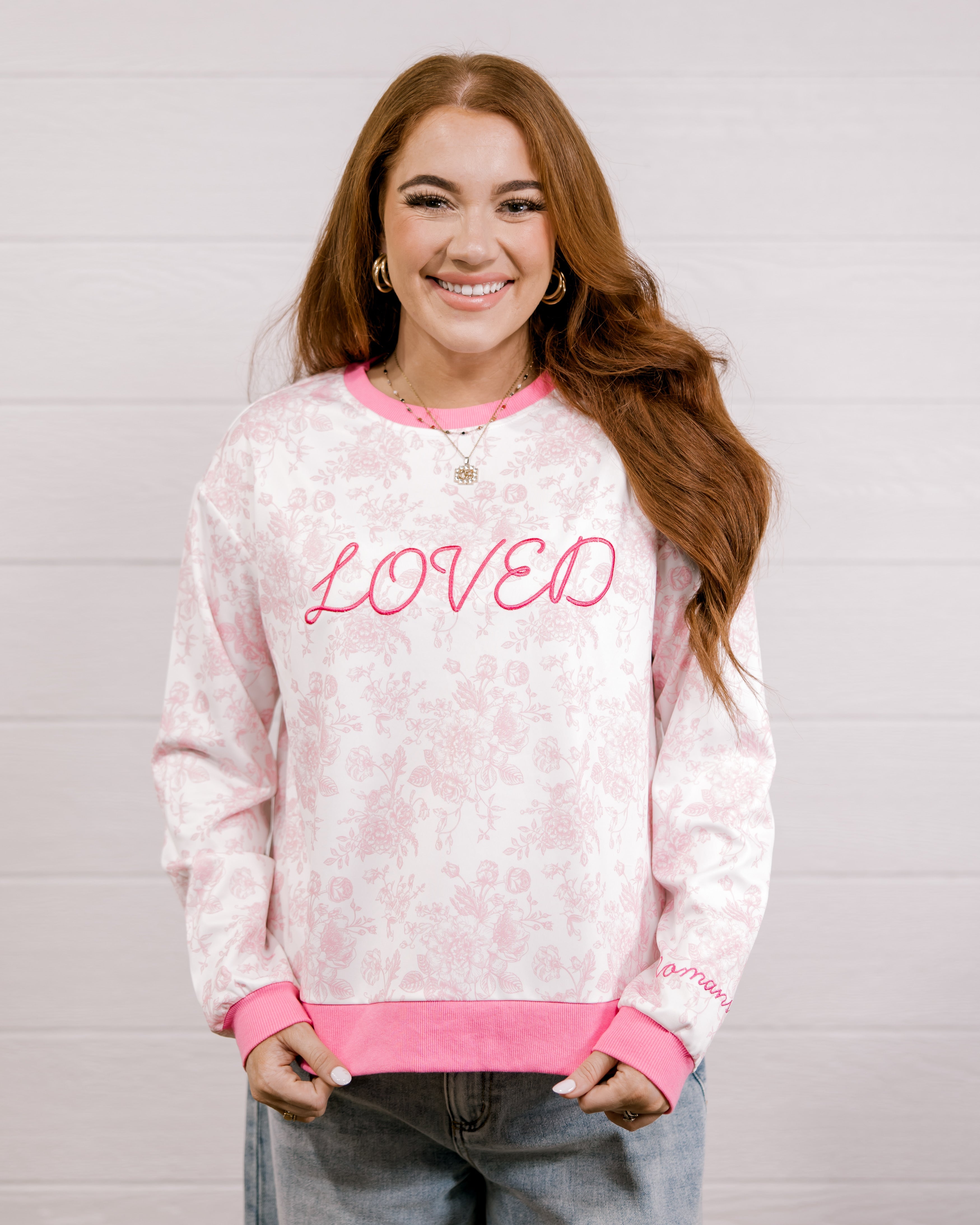 Woman wearing a pink and white patterned sweatshirt with 'LOVED' embroidered in pink standing against a white background.
