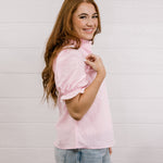 Side view of woman wearing a pink & white gingham blouse with an elbow sleeve and lace detail on trim against a white wall.