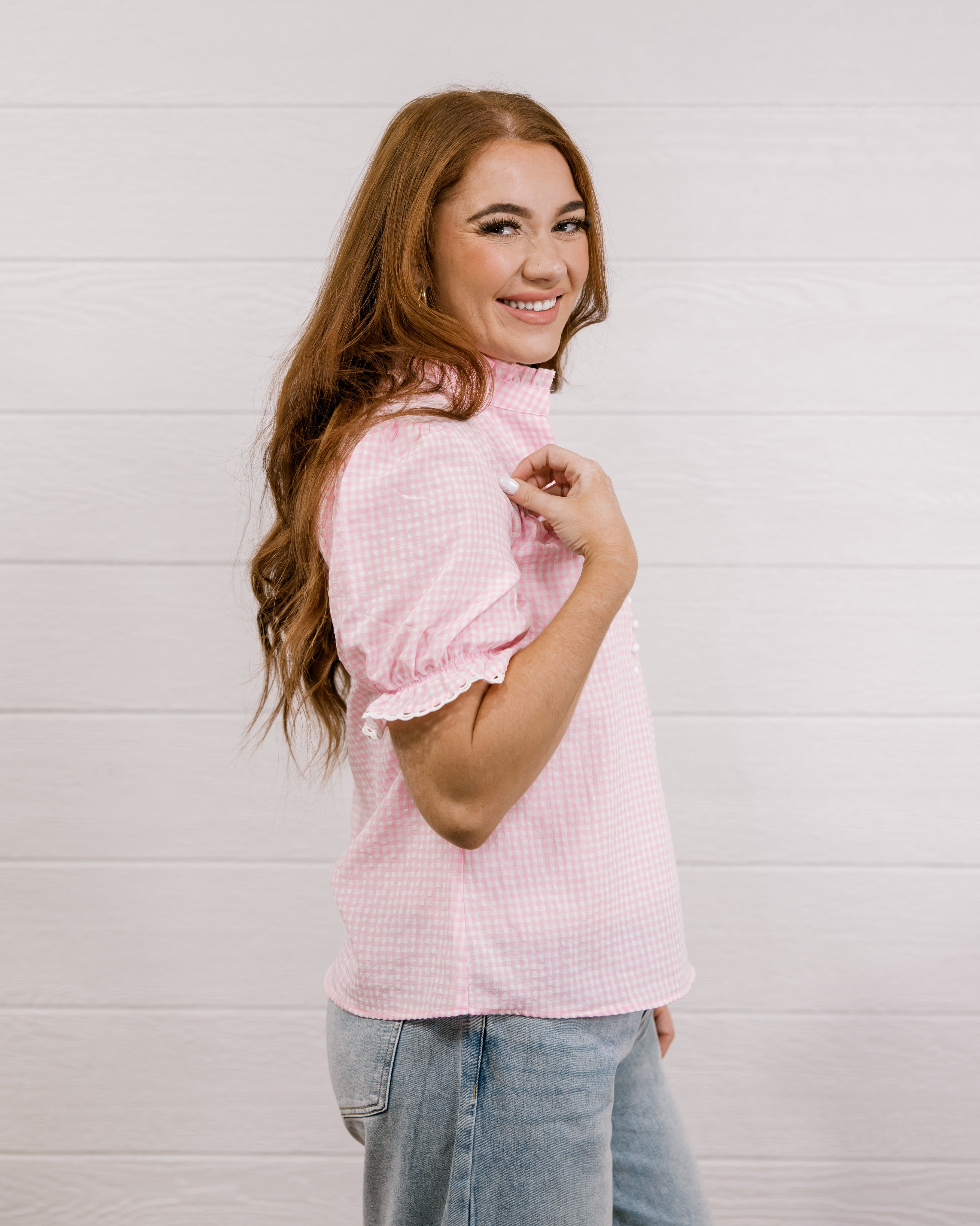Side view of woman wearing a pink & white gingham blouse with an elbow sleeve and lace detail on trim against a white wall.