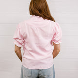Back view of woman wearing a pink & white gingham blouse with an elbow sleeve and lace detail on trim against a white wall.