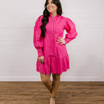 Woman wearing a bright pink eyelet dress standing on a wooden floor with a white wall background