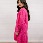 Woman wearing a bright eyelet pink dress against a white wall.