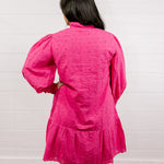 Woman wearing a bright pink eyelet dress with puffed sleeves against a white wall.