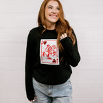 Woman wearing a black sweater with a playing card graphic design featuring a red cowgirl and sequin hearts against a white background. 