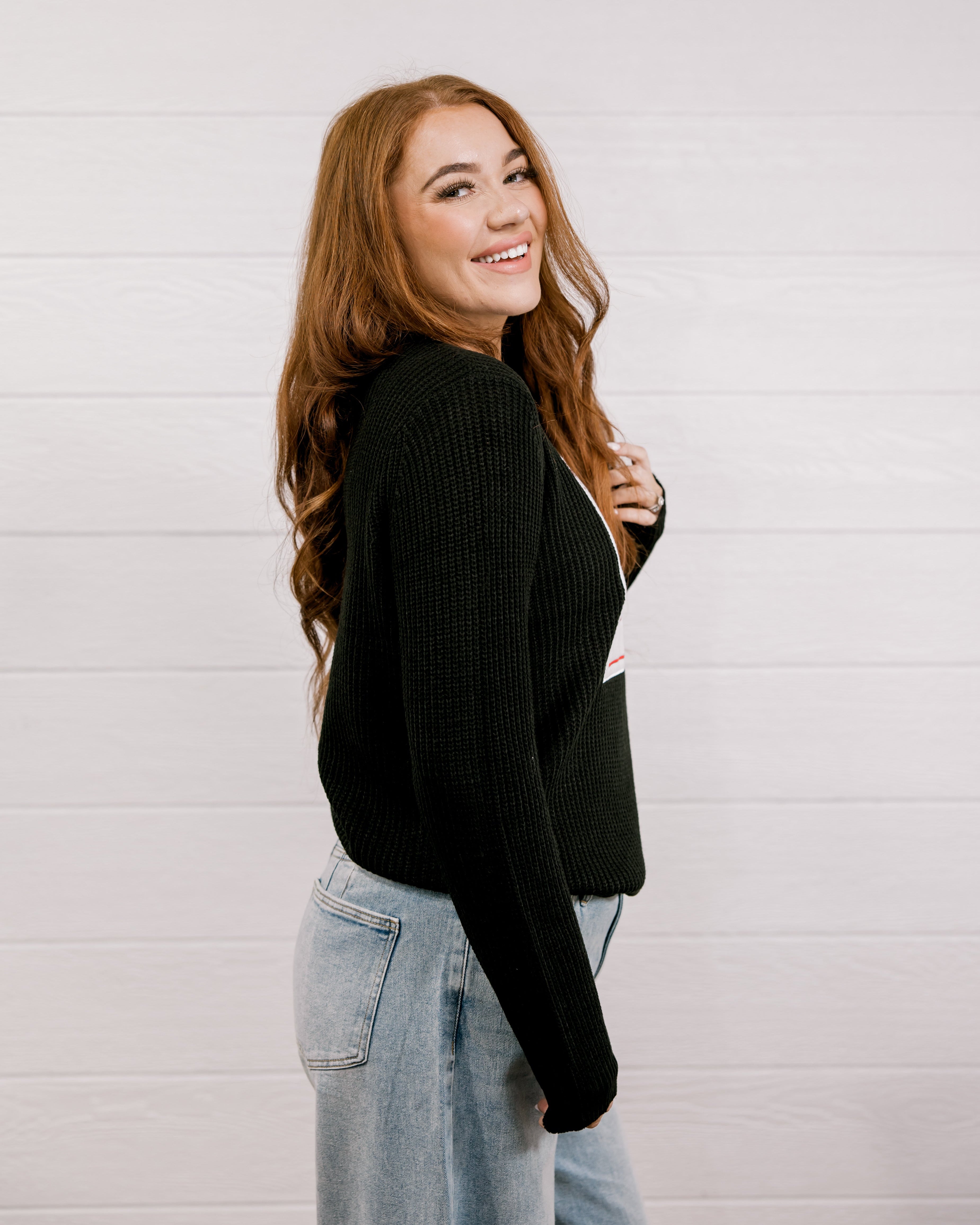Side view of woman wearing a black sweater with a playing card graphic design featuring a red cowgirl and sequin hearts against a white background. 